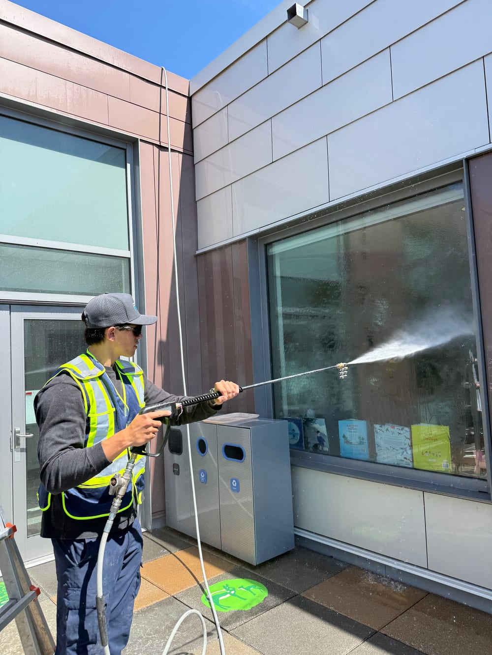 A Flat Mate technician in high-visibility gear soft washing the upper facade and glazing of a commercial building with a pole-fed system